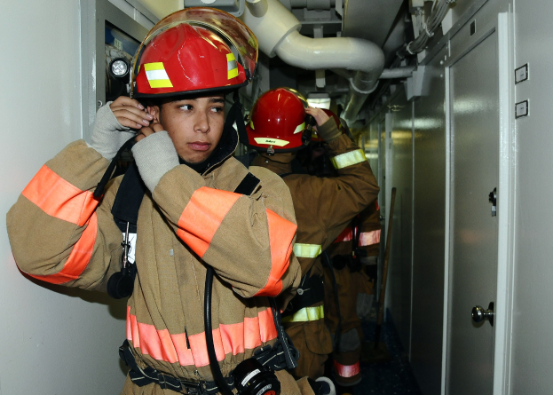 Gruppe von Feuerwehrleuten in Uniform, die nebeneinander in einem Trainingsraum mit einer Tür auf der rechten Seite und einer Wand auf der linken Seite stehen. Im Hintergrund sind Rohre und andere Gegenstände zu sehen.