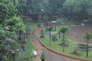 Ein grüner Park mit Bäumen, Pflanzen, Gras, Laternen, Bänken und Skulpturen sowie einem Gebäude im Hintergrund.