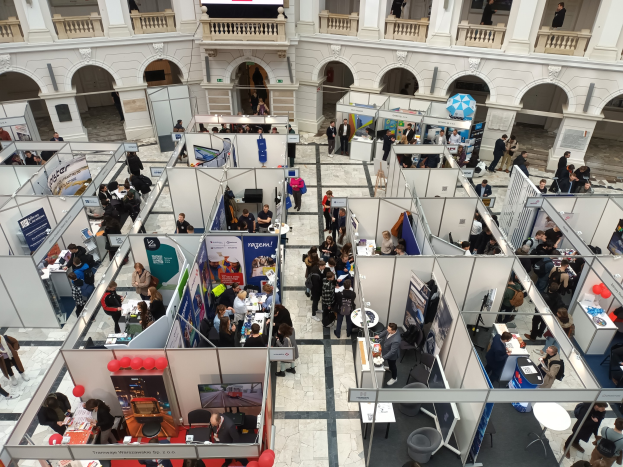 Große Gruppe von Menschen auf einer internationalen Fachmesse mit Ständen, Bannern, Tischen, Stühlen und Ballons in einem Raum mit architektonischen Bögen und Säulen.