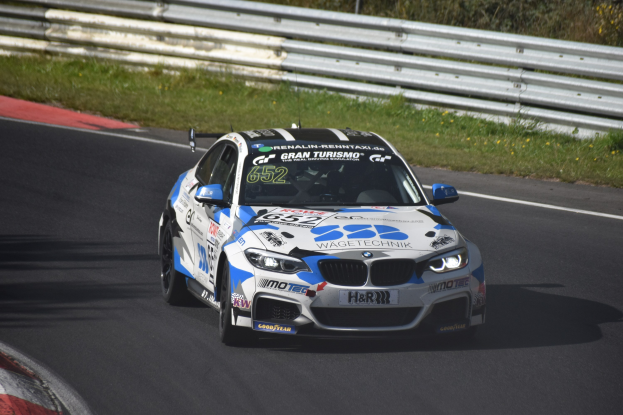Ein BMW M4 DTM Rennwagen mit Text und Zahlen auf seiner Karosserie, der auf einer Rennstrecke fährt, umgeben von Gras, einem Zaun und Pflanzen, mit einem sichtbaren Rad.