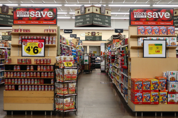 Innensicht Eines Kiosks Mit Verschiedenen Lebensmitteln Auf Regalen Und Dem Boden, Beleuchtet Von Deckenlampen, Und Mit Informationsschildern Mit Text Und Zahlen.