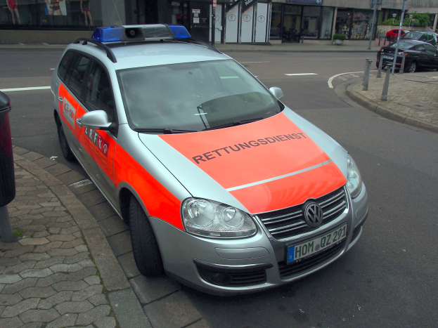 Polizeiauto am Straßenrand geparkt mit einem Müllcontainer auf dem Gehweg zur Linken, Gebäude und Bäume im Hintergrund.