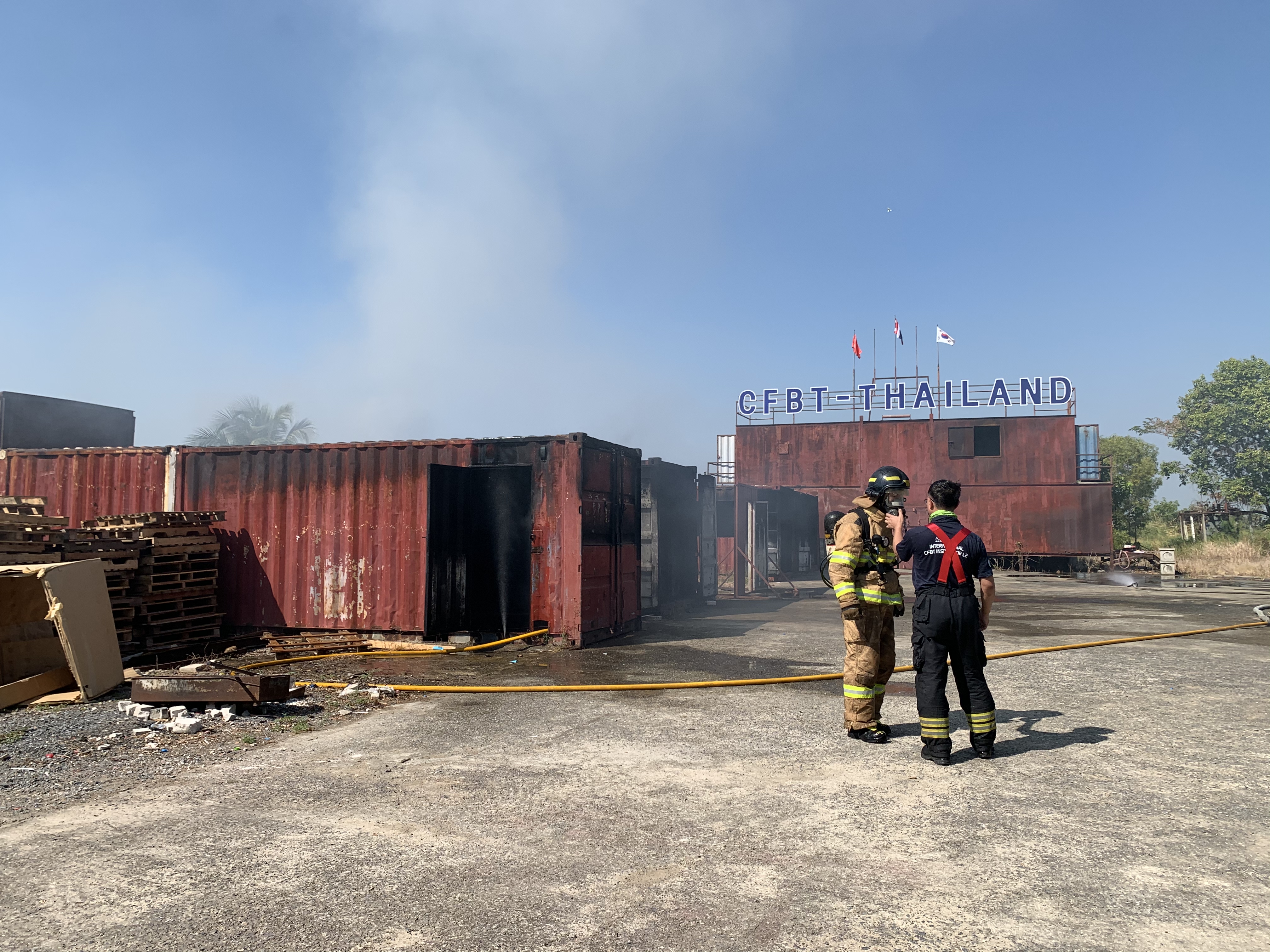 Zwei Feuerwehrleute in Helmen vor einem qualmenden Gebäude mit verstreuten Containern und Holzgegenständen unter einem klaren blauen Himmel mit Bäumen im Hintergrund.