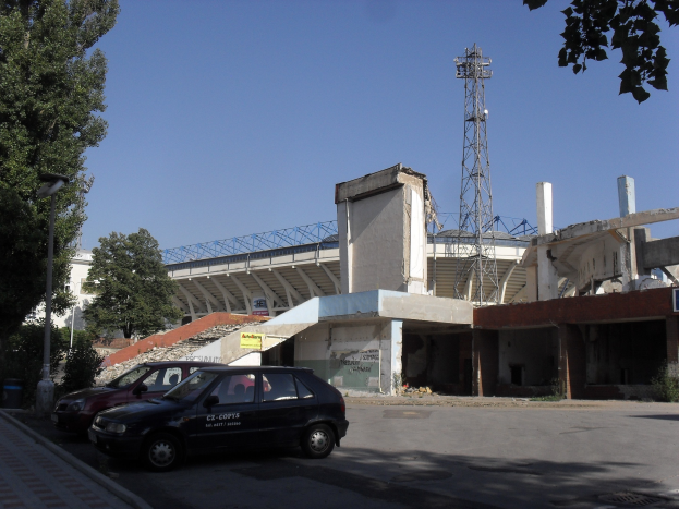 Alter Stadion mit parkenden Autos davor, umgeben von Gebäuden, Bäumen, Pfählen und einem Turm unter einem sichtbaren Himmel.