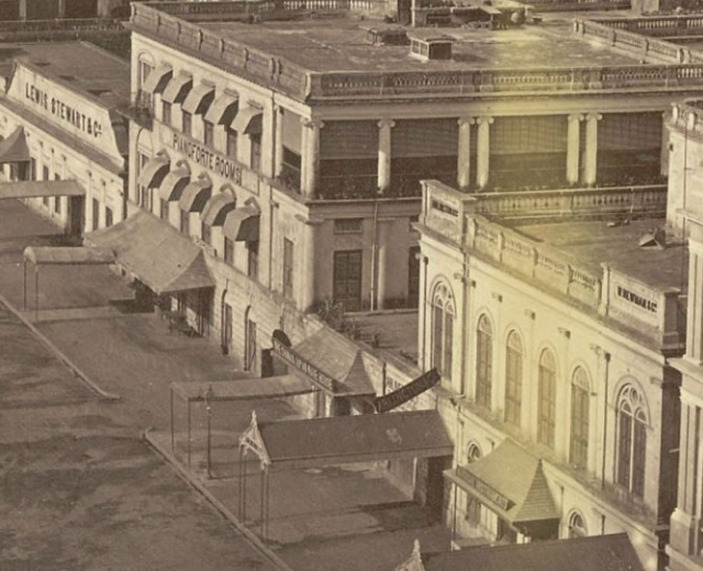 Altes Schwarz-Wei├č-Foto einer Stadtstra├če mit Geb├Ąuden, Fenstern, S├Ąulen, Namensschildern, Schuppen und Masten.