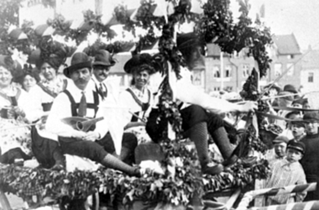 Eine Gruppe von Menschen in traditioneller bayrischer Tracht sitzt auf einem mit Laub geschmückten Festwagen bei einem Umzug, einige spielen Musikinstrumente, mit Gebäuden und einem klaren Himmel im Hintergrund, dargestellt in Schwarz-Weiß.