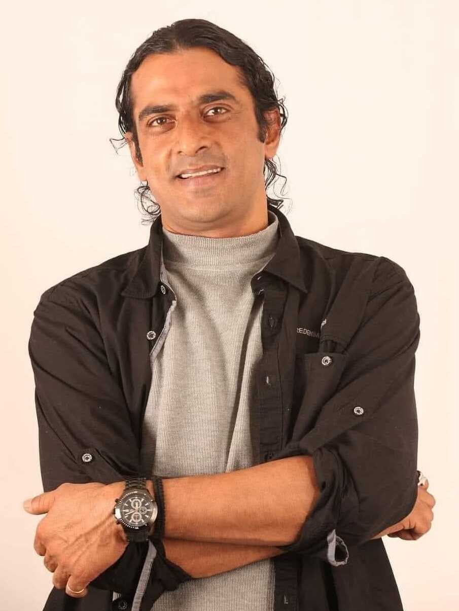 Ein Mann in einem schwarzen und aschfarbenen Kleid mit überschlagenen Armen, der eine Uhr tr├Ągt und gegen einen weißen Hintergrund steht, der tief nachdenklich erscheint.