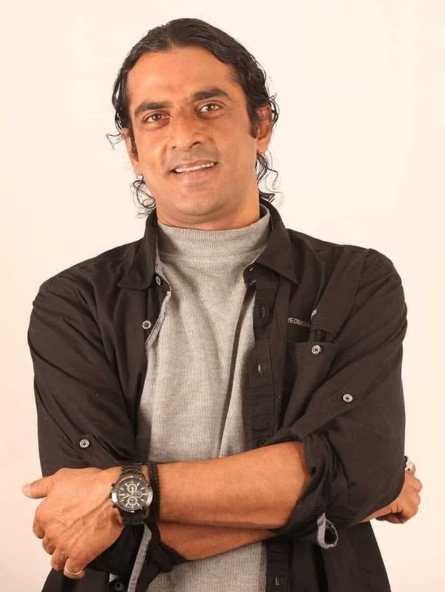 Ein Mann in einem schwarzen und aschfarbenen Kleid mit überschlagenen Armen, der eine Uhr tr├Ągt und gegen einen weißen Hintergrund steht, der tief nachdenklich erscheint.