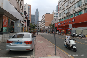 Ein Mann fährt mit einem Roller eine Straße in der Stadt entlang, gesäumt von hohen Gebäuden, mit Fußgängern auf den Bürgersteigen und anderen Fahrzeugen auf der Straße, unter einem klaren blauen Himmel mit Bäumen im Hintergrund.