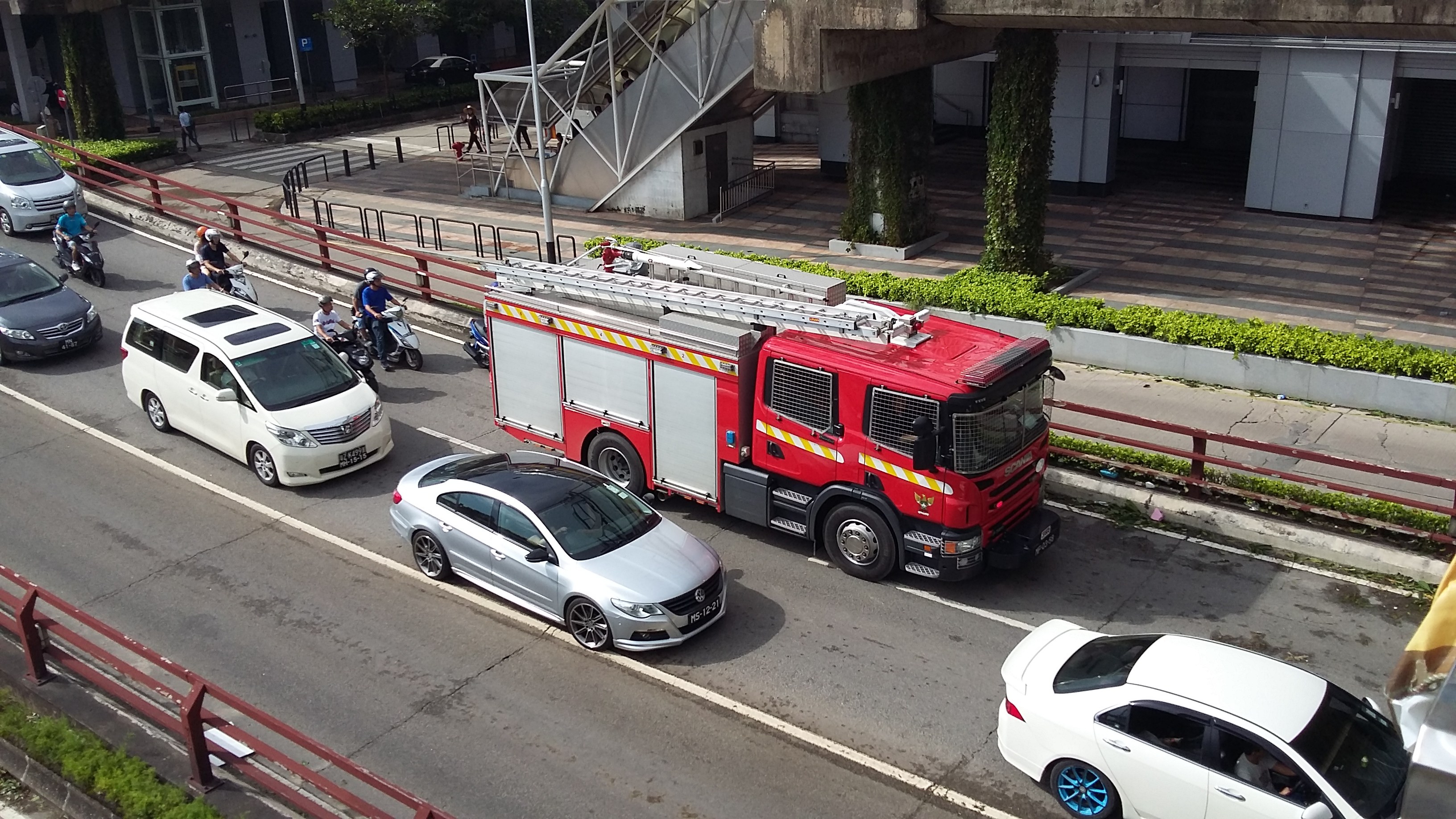 Feuerwehrfahrzeug fährt auf einer Straße mit Autos, Motorrädern und anderen Fahrzeugen; Gebäude und Bäume sind im Hintergrund zu sehen.