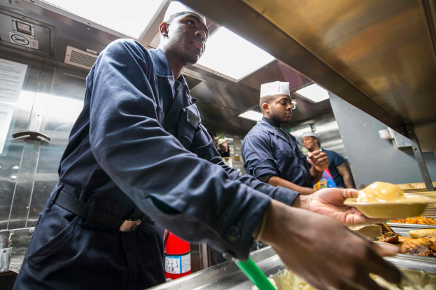 Eine Gruppe von M├Ąnnern steht in einer K├╝che, einer h├Ąlt einen Teller mit Essen, mit Tabletts voller Essen auf dem Tisch, einem Feuerl├Âscher und Papieren an der Wand im Hintergrund.