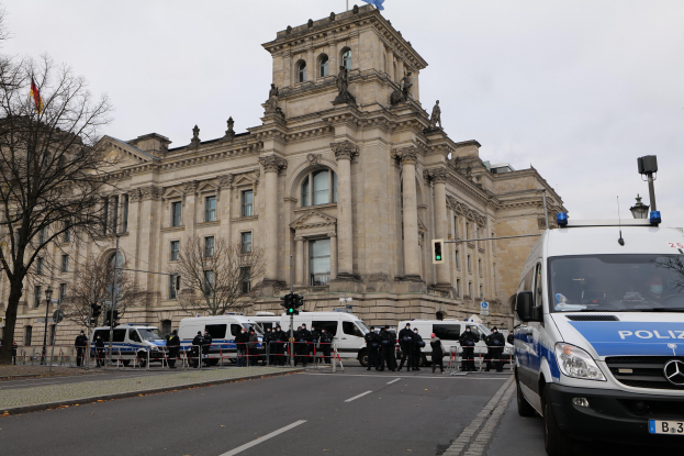 Gruppe von Polizeibeamten vor dem Reichstaggebäude in Berlin, Deutschland, mit Fahrzeugen, einem Zaun, Verkehrsampeln, Laternenmasten, Bäumen und Flaggen im Hintergrund.