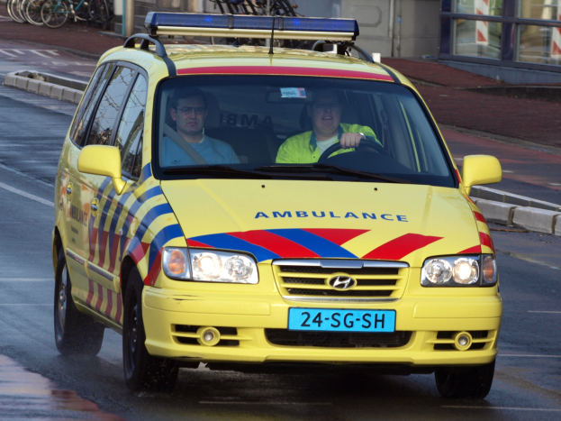 Gelber Krankenwagen fährt auf der Straße mit zwei Personen darin, Fahrräder sind auf dem Gehweg geparkt und im Hintergrund ein Gebäude mit Glasfenstern.
