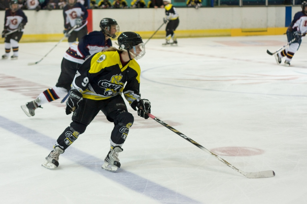 Menschen beim Schneehockey mit Hockey-Schlägern und Helmen, mit anderen Menschen und einer Wand im Hintergrund.