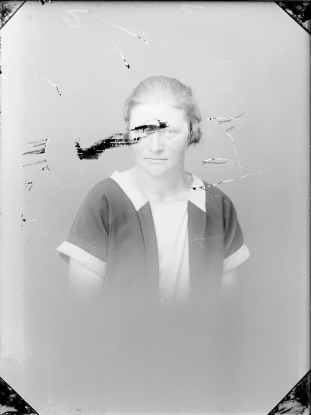 Ein Schwarz-Weiß-Foto einer Frau mit einem zerbrochenen Glasfenster im Hintergrund, ihr Gesicht ist sichtbar, Blick direkt und leicht geöffnete Lippen, Haar zurückgebunden, Hände vor der Brust verschränkt.