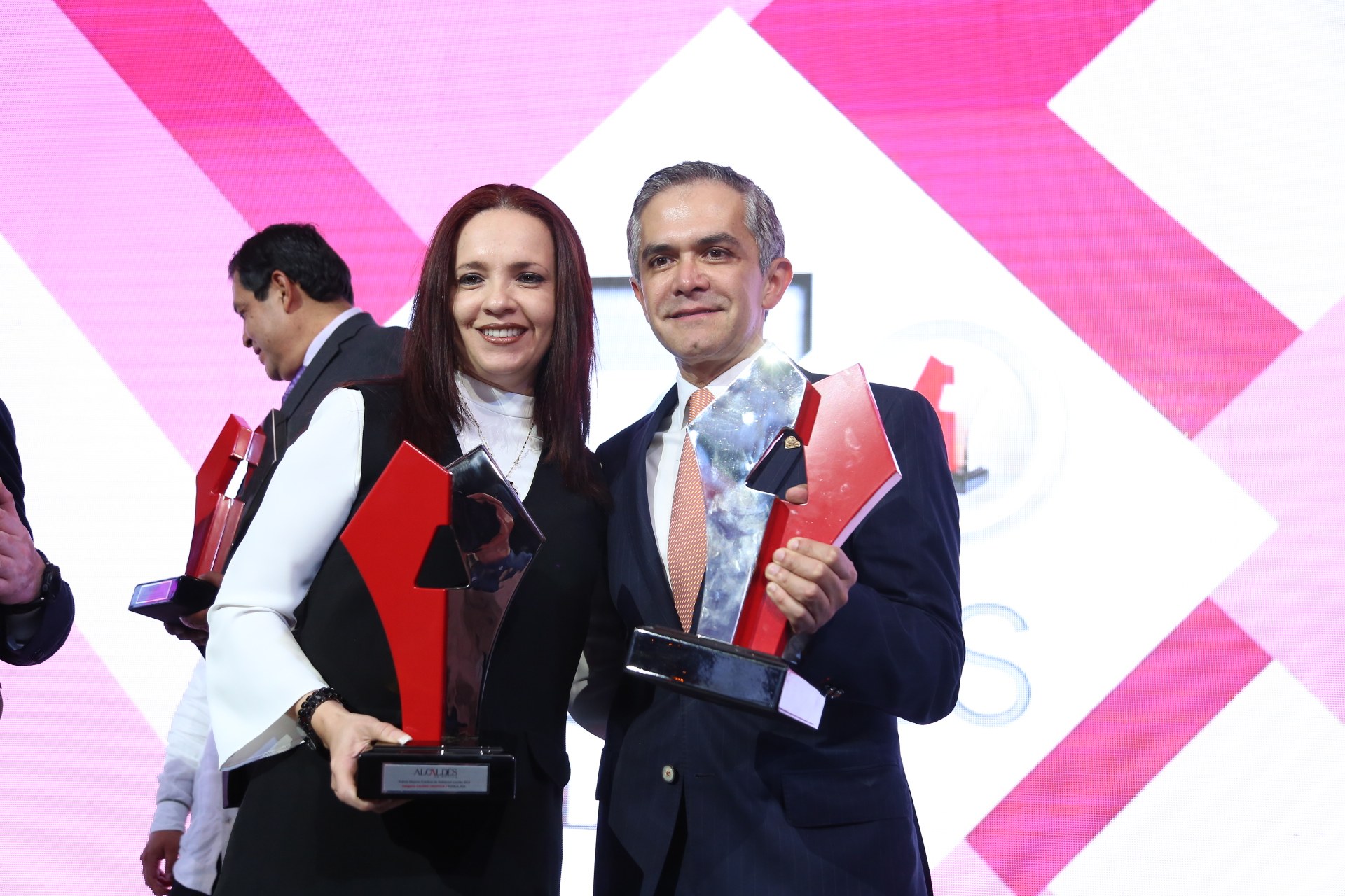 Zwei Personen auf einer Bühne, die lächeln und Trophäen halten, mit einem Bildschirm im Hintergrund.