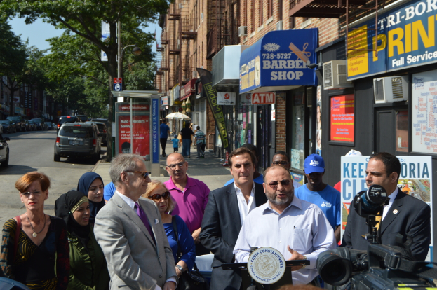 Eine Gruppe von Menschen mit Kameras, eine Person in einem weißen Hemd mit einem Mikrofon und einer Schutzbrille, mit parkenden Autos, Geschäften, Gebäuden, Bäumen, Straßenlaternen und Passanten im Hintergrund bei klarem Himmel.