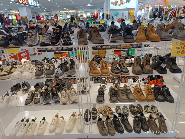 Ein Schuhgeschäfts-Innenraum mit verschiedenen Schuharten auf Ständern, Preisetiketten sichtbar, Menschen im Hintergrund, Kleiderauslagen, Deckenlampen, ein Bildschirm und Texttafeln.