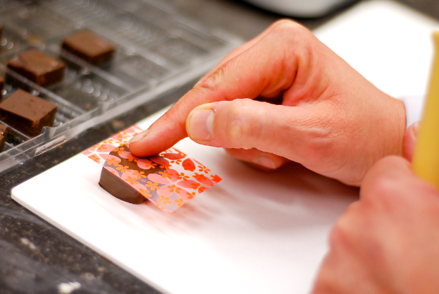 Eine Hand legt eine dekorative Abdeckung auf eine Schokolade, mit mehr Schokoladen auf einem Tablett links und einem Tisch darunter.
