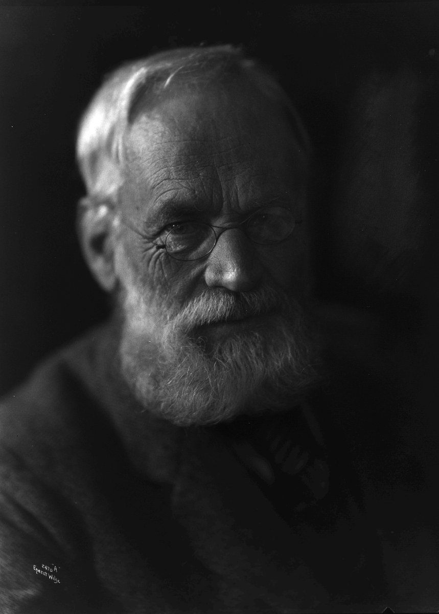 Alter Mann mit Vollbart und Brille, der einen dunklen Anzug trägt und ernst in die Ferne schaut.