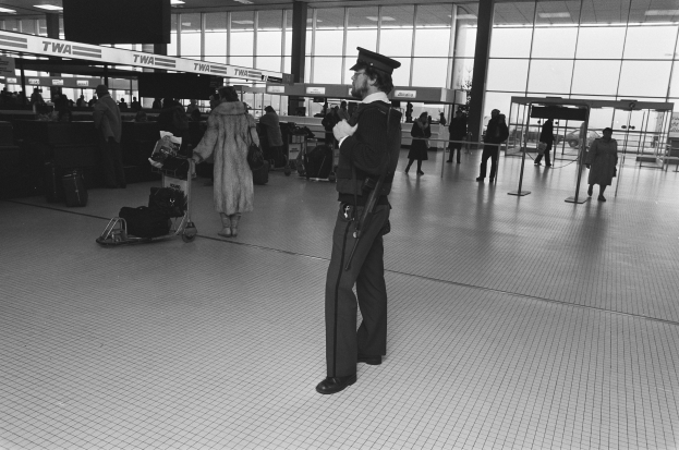 Polizeibeamter in Uniform mit Mütze, der eine Waffe hält und in einem Flughafen-Terminal steht, umgeben von Menschen, Gepäckwagen und verstreuten Taschen, mit Hintergrund-Elementen wie Pfosten, Texttafeln, Deckenleuchten und Glaswänden.