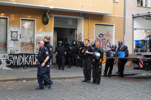 Eine Gruppe von Polizisten, einige mit Helmen und Waffen, steht und geht vor einem mit Graffiti beschmierten Gebäude, mit einem Fahrzeug und verschiedenen Gegenständen im Hintergrund.