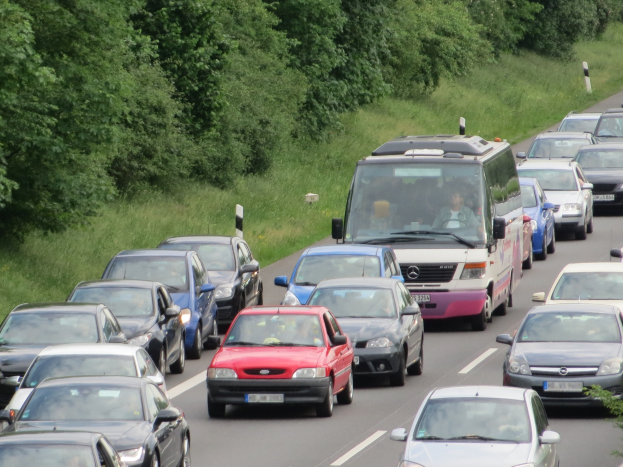 Ein Stau auf einer Autobahn mit zahlreichen Autos und einem Van, mit Menschen in den Fahrzeugen und Bäumen und Gras im Hintergrund.