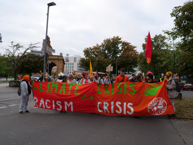 Eine Gruppe von Menschen marschiert eine baumbewachsene Straße entlang und hält ein Banner mit der Aufschrift "Klimakrise ist eine Krise", mit Fahrzeugen und Gebäuden im Hintergrund unter einem klaren blauen Himmel.