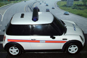 Ein weißes Polizeifahrzeug steht im Vordergrund mit mehreren fahrenden Autos auf der Straße dahinter, umgeben von Gras und Bäumen in einer ländlichen Umgebung.