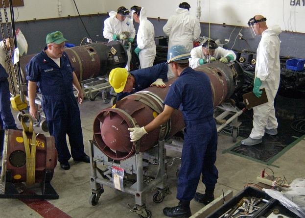 Männer in Helmen und Handschuhen beim Entschärfen einer großen Bombe in einer Fabrik mit Maschinen und Wandtext.