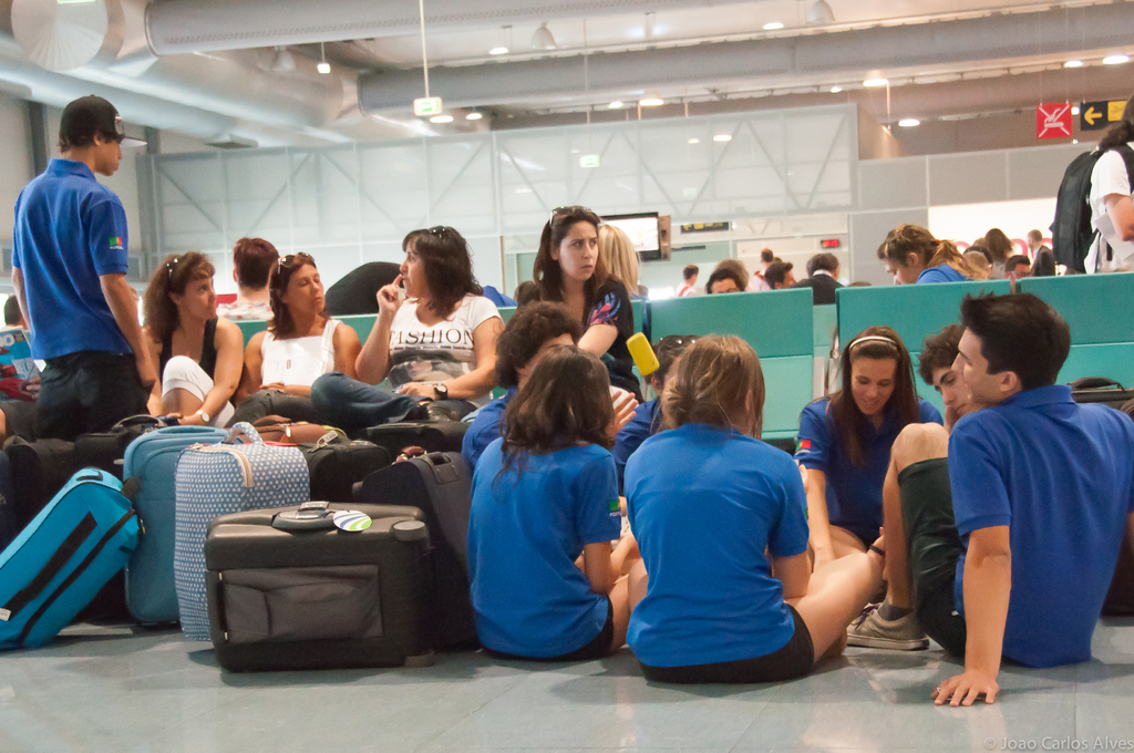 Ein belebter Flughafeninnenraum mit vielen Menschen, die auf Flüge warten, Gepäck auf der linken Seite und Glasdoors mit Richtungsschildern im Hintergrund.