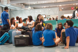 Ein belebter Flughafeninnenraum mit vielen Menschen, die auf Flüge warten, Gepäck auf der linken Seite und Glasdoors mit Richtungsschildern im Hintergrund.