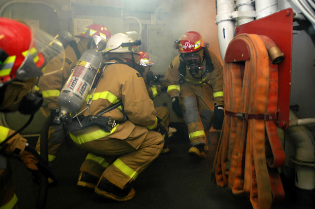 Feuerwehrleute in Schutzausrüstung mit Rauch, die neben Rohren und einer Wand stehen.
