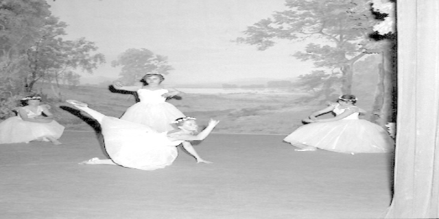 Vier junge Balletttänzerinnen in weißen Kleidern tanzen auf einer Bühne mit einer gemalten Kulisse aus Bäumen und Himmel, in Schwarz-Weiß.