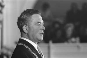 Ein Mann im Anzug und Krawatte steht an einem Podium mit einem Mikrofon und spricht zu einer unscharfen Menge, der Anschein eines Politikers.