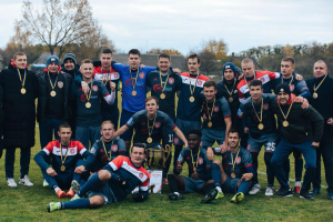 Eine Gruppe von Männern in Medaillen steht auf einem Fussballfeld und hält einen Pokal, umgeben von Bäumen unter einem klaren blauen Himmel.