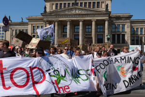 Eine Gruppe von Menschen mit Schildern und Plakaten vor einem Gebäude mit Fenstern, Säulen, Statuen und einer Tafel, die an einer Klimademonstration in Berlin teilnehmen, mit dem Himmel im Hintergrund.