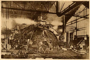 Ein Schwarz-Weiß-Foto eines Fabrikinneren mit Arbeitern, die eine große Maschine bedienen, umgeben von Leitern und Stangen, mit dem Text "Cadillac Steel Works" unten.