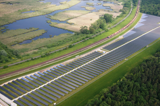 Luftaufnahme eines großen Solarparks in einem Feld mit Solarpanelen, umgeben von Bäumen, Gras, Pflanzen und Wasser, mit einem Zug auf einer nahen Eisenbahnstrecke.