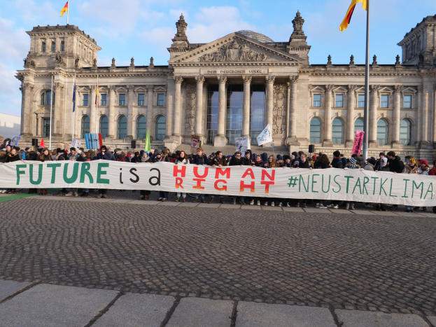 Eine Gruppe von Menschen hält ein Transparent mit der Aufschrift "Zukunft ist ein Mensch" vor dem Reichstagsgebäude in Berlin, Deutschland, das mit Fenstern, Säulen, Bögen und Statuen verziert ist und von Fahnen mit Stangen umgeben ist, unter einem bewölkten Himmel.