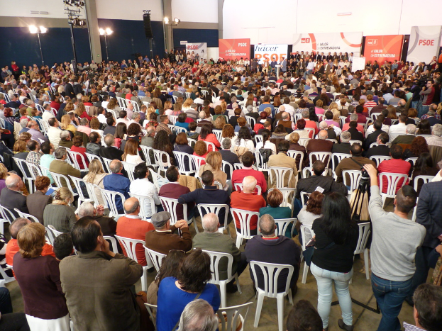 Ein voller Stadion mit vielen sitzenden und stehenden Menschen, eine Bühne mit zwei Rednern an einem Podium und mehrere Banner und Lichter in der Nähe.