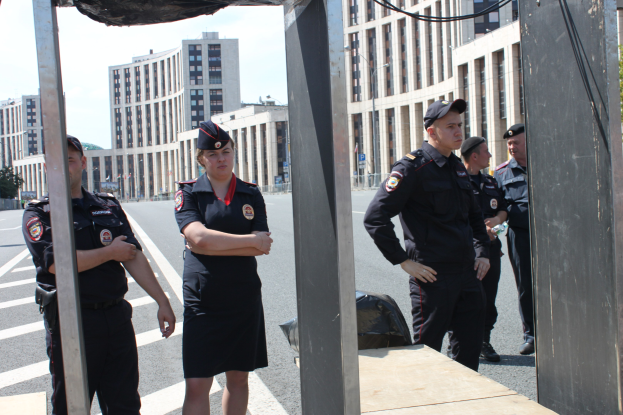 Gruppe von Polizisten in Uniform auf einer Straße mit Masten und Drähten im Vordergrund, Gebäuden, Bäumen und einem klaren blauen Himmel im Hintergrund.