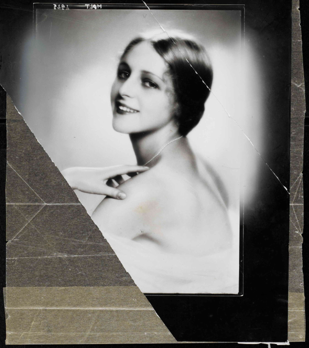 Ein Schwarz-Weiß-Porträt einer Frau in einem Kleid im Stil der 1920er Jahre mit ihrem Haar zu einem ordentlichen Knoten frisiert, die warm in die Kamera lächelt.