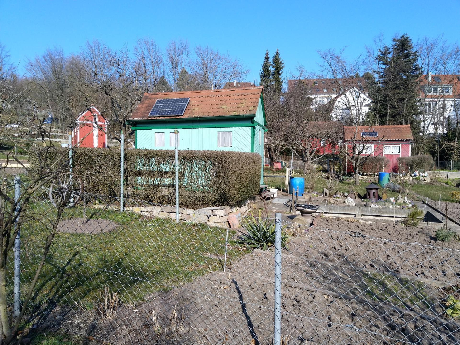 Kleines grünes Haus mit Solarpanelen auf dem Dach, umgeben von Maschendrahtzaun, umgeben von Bäumen, Pflanzen und Gras unter einem klaren blauen Himmel.