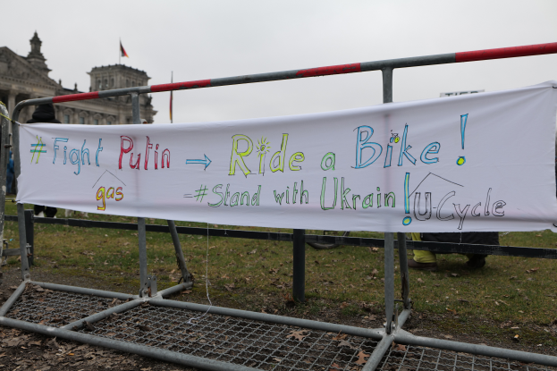 Eine Gruppe von Menschen hält ein Banner mit der Aufschrift "Kämpfe gegen Putin, Fahrrad fahren, Stehe mit der ukrainischen U-Cycle" auf dem Boden neben einem Metallzaun, trockene Blätter, ein Gebäude mit Fenstern, ein Fahnenmast mit einer Flagge, unter einem bewölkten Himmel.