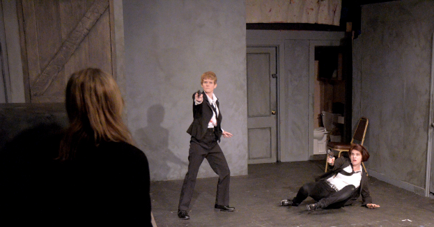 Zwei Frauen in schwarzen Anzügen, eine sitzend auf dem Boden und die andere stehend, beide mit Waffen in den Händen, sowie eine dritte Frau in der Nähe, in einem Raum mit einem Stuhl, einer Toilette und einer hölzernen Tür.