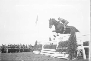 Schwarzes und weißes Foto eines Pferdes und Reiters, die über ein Hindernis springen, bei den 1953 Royal Ascot Horse Trials, mit Zuschauern auf der linken Seite, einer Fahne im Hintergrund und Gras am Boden.