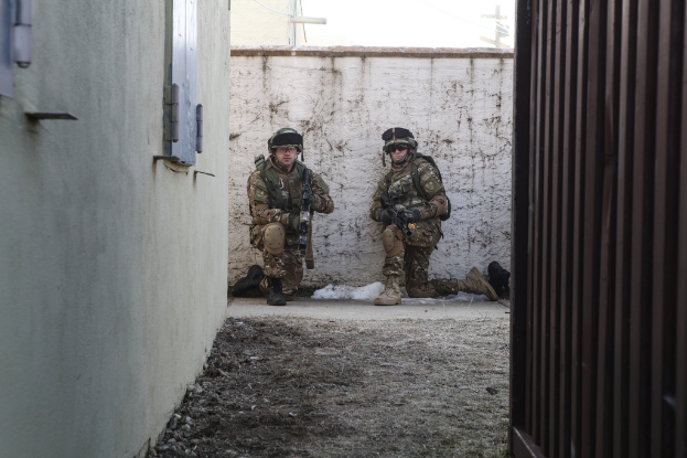 Eine Szene mit einer Wand und Fenster auf der linken Seite, einer Eisenwand auf der rechten Seite, zwei Soldaten und Erde in der Mitte und Gebäuden, einem Strommast und einer weiteren Wand im Hintergrund.