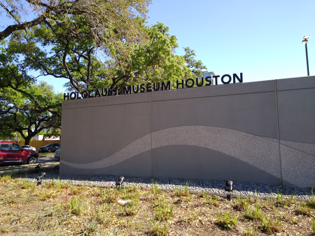 Das Holocaust-Museum in Houston, Texas, mit einer textbedeckten Wand, Steinen, Gras, Pflanzen, einer Baumgruppe, Fahrzeugen auf der Straße und einem Wolkenhimmel.