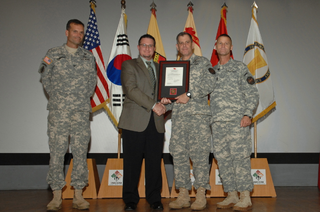 Vier Männer stehen auf einer Bühne: drei in Militäruniformen und einer in Blazer, Krawatte und Hemd, die Hände schütteln und einen Preis überreichen, mit sechs Flaggen auf Ständen und einer weißen Wand im Hintergrund.